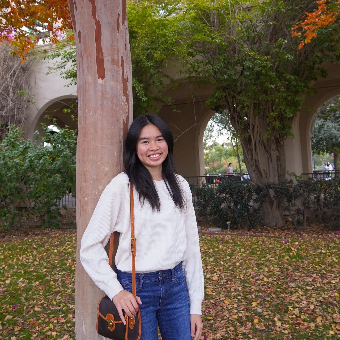 woman with black hair leaning on a tree wearing a white sweater